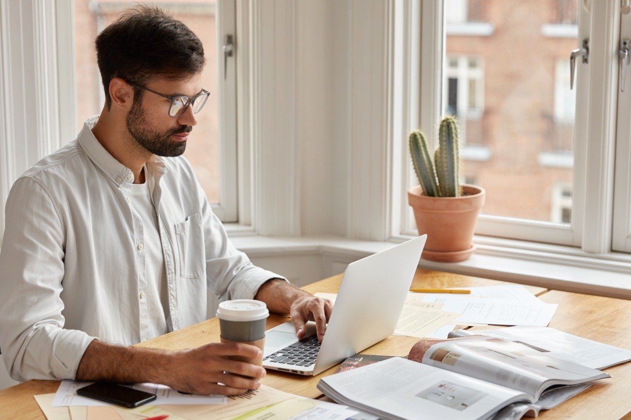 Image of concentrated unshaven businessman watches important webinar or online conference, holds takeaway coffee, wears spectacles for good vision, surrounded with journal and paper documents