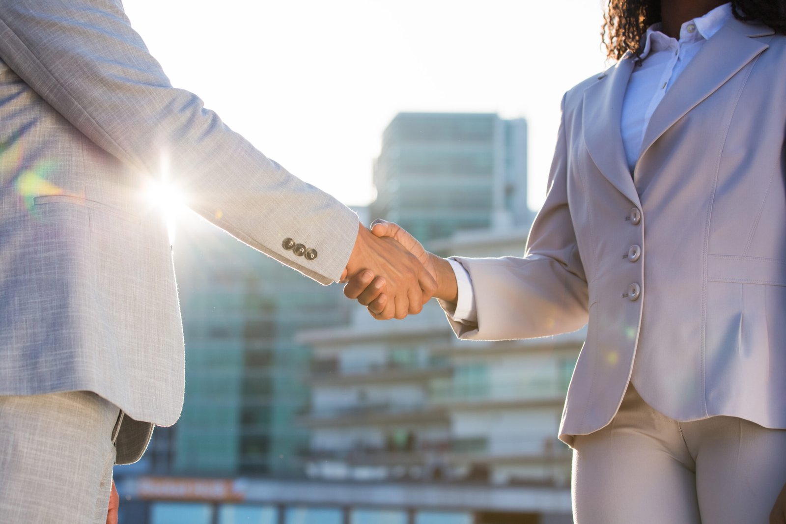 Business partners meeting in city in early morning. Business man and woman shaking hands in sunlight. Urban buildings in background. Corporate meeting concept