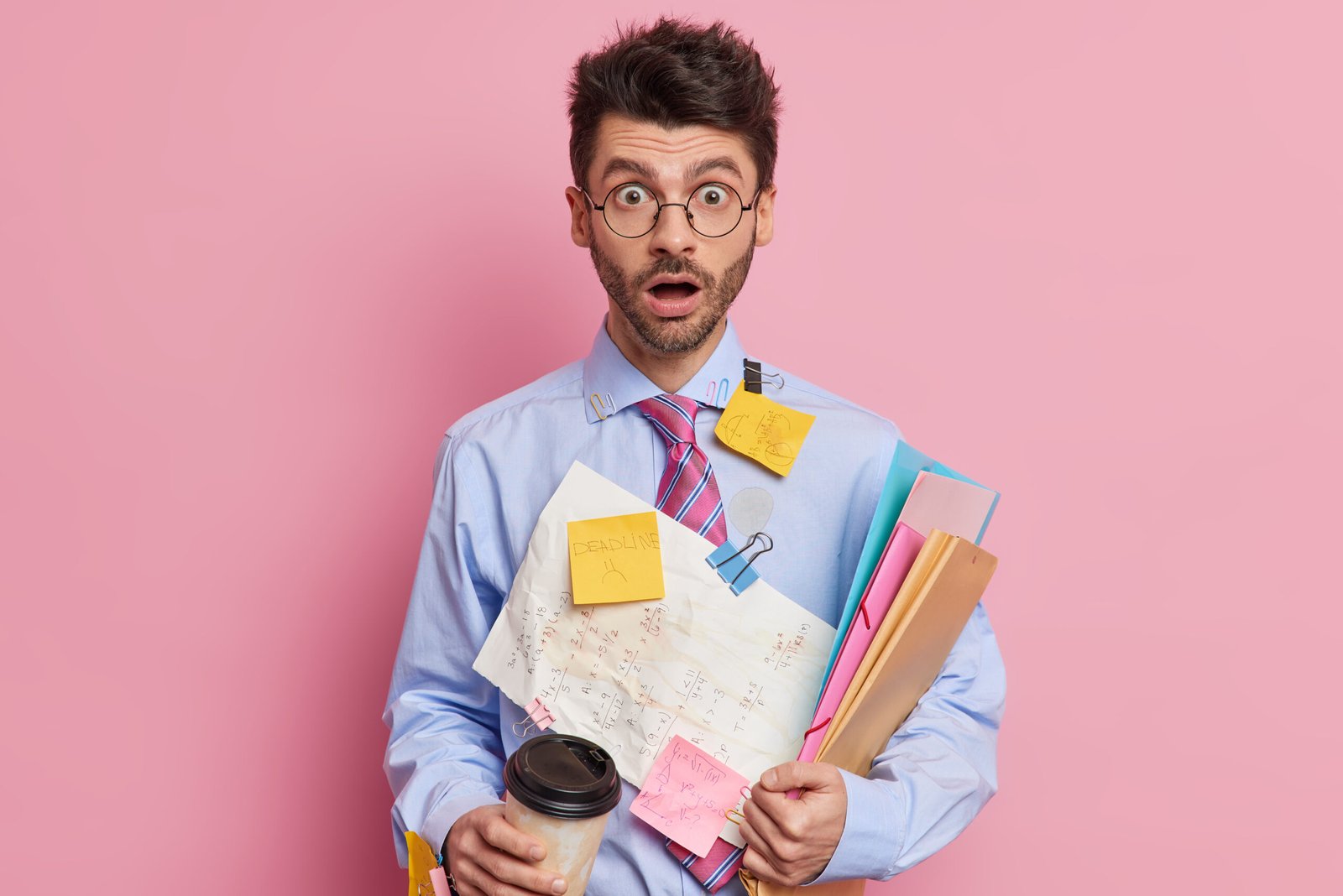 Indoor shot of shocked skilled male student stares with eyes popped out and jaw dropped works on project or prepares for exams writes down information on papers to remember drinks coffee to go