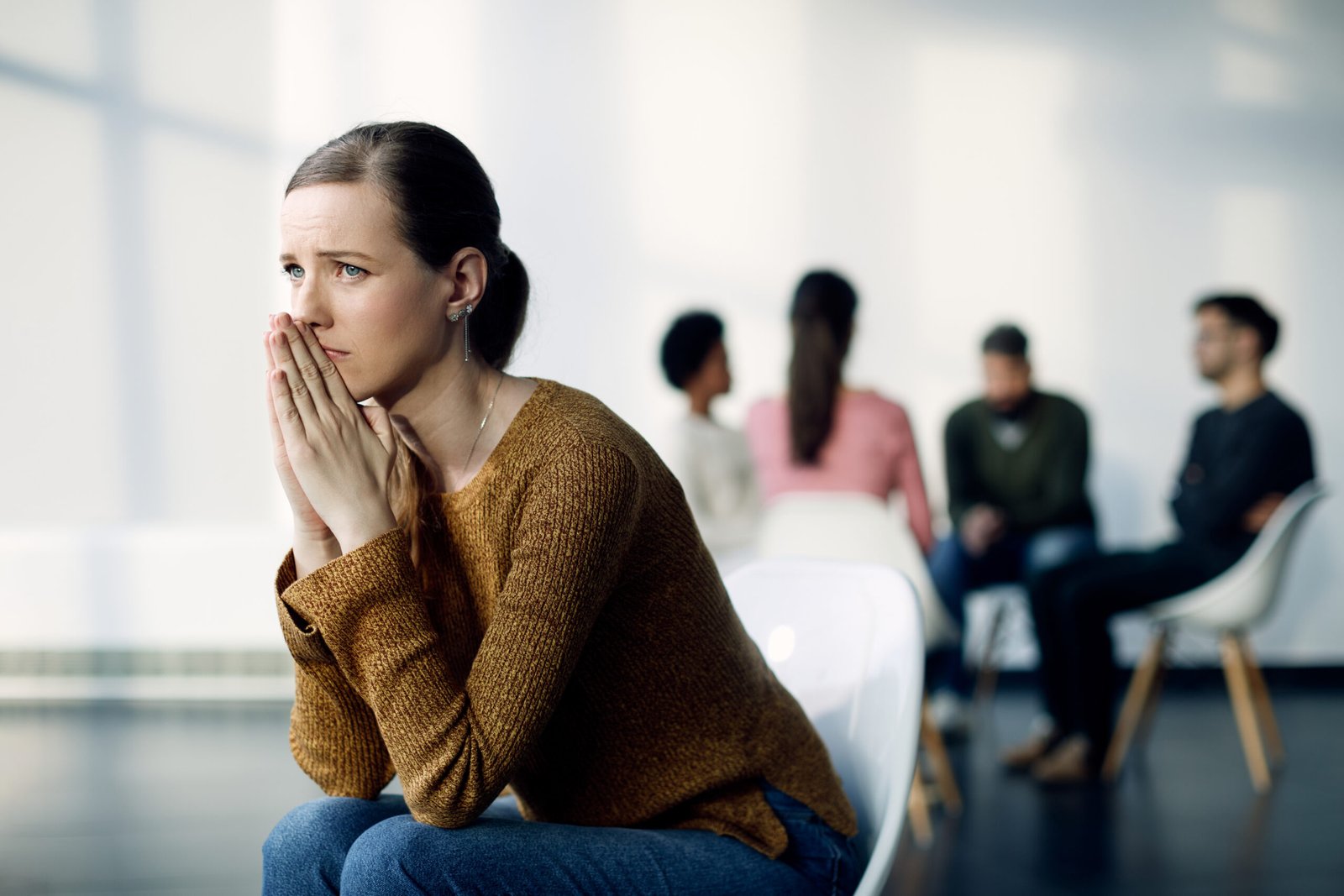 Young woman feeling sad while sitting apart from group therapy participants at community center.